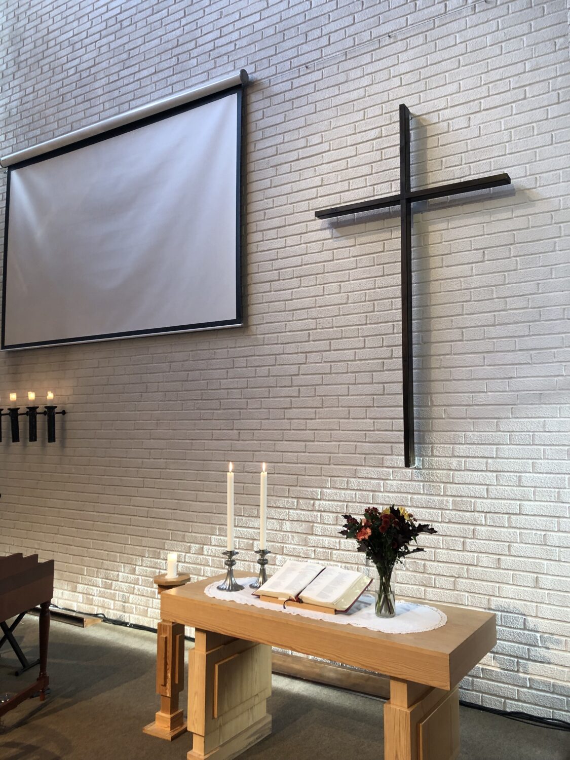Altarbord med två stearinljus, en bibel, blommor och ett kors på väggen.